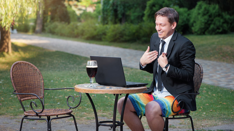 man in a video conference call wearing swimming trunks, shirt, tie, and suit jacket with a beer behind the laptop