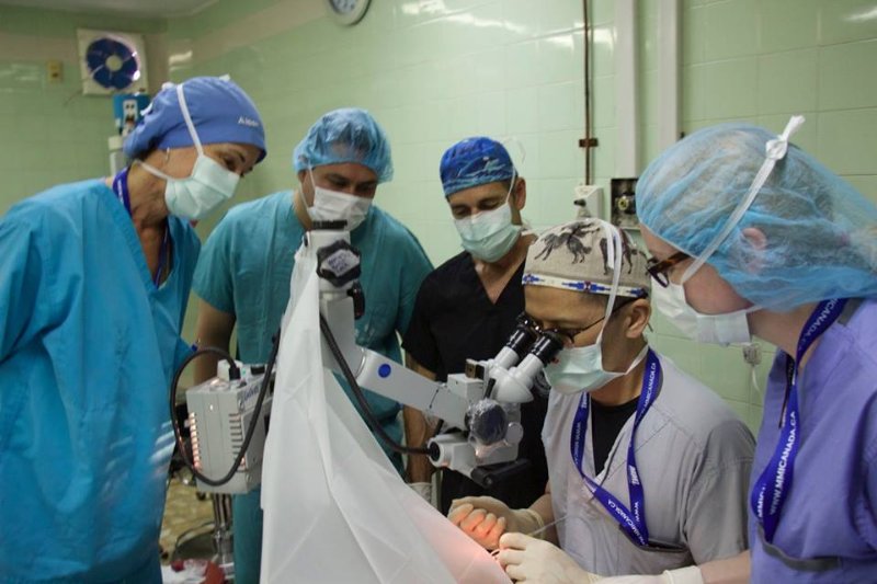 a person looking into a microscope surrounded by four people in surgical gowns and masks