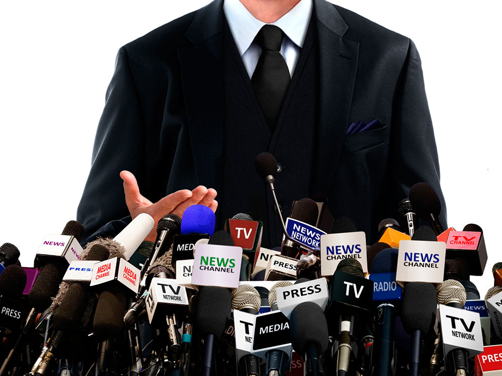 man talking in front of a whole lot of media microphones